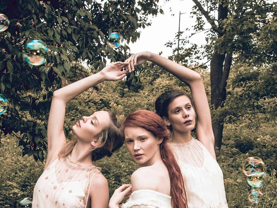 Three fashion models in pale dresses posing beneath raised arms with bubbles floating around them.