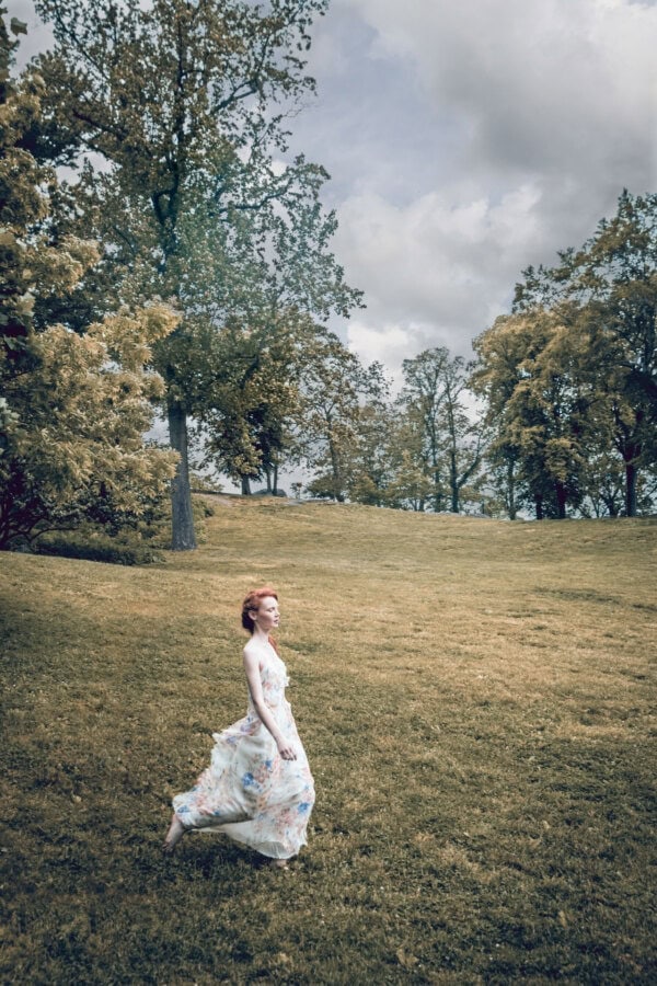 Red-haired fashion model in a flowing floral dress walking alone across a wide grassy hillside.