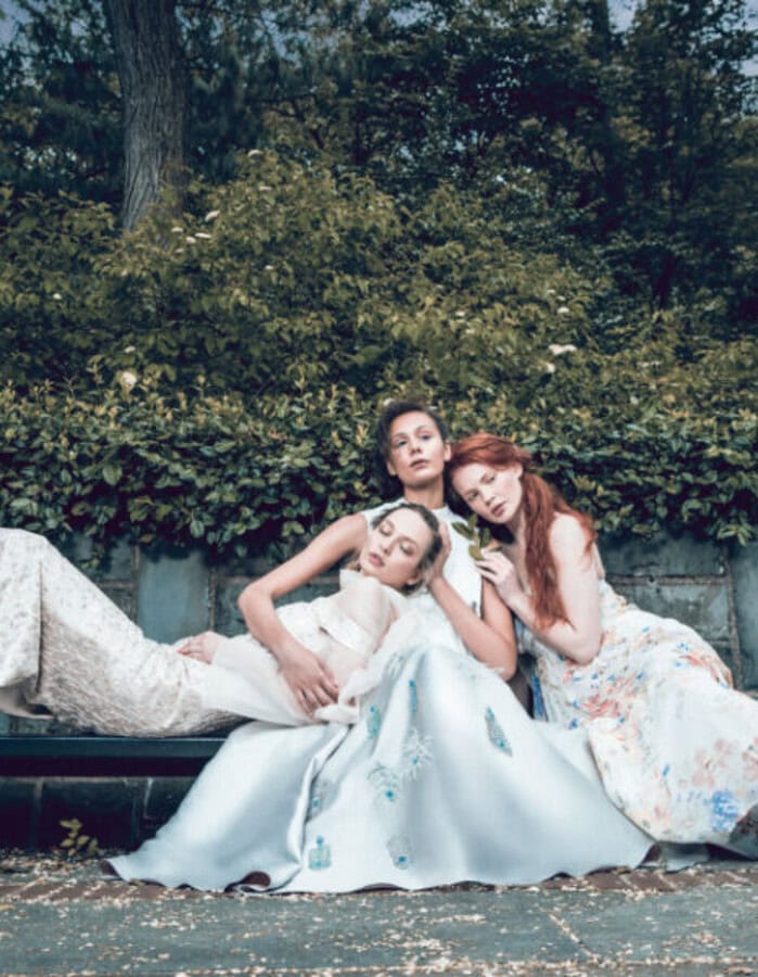 Three fashion models in flowing pale dresses reclining together on a park bench beneath dense greenery.