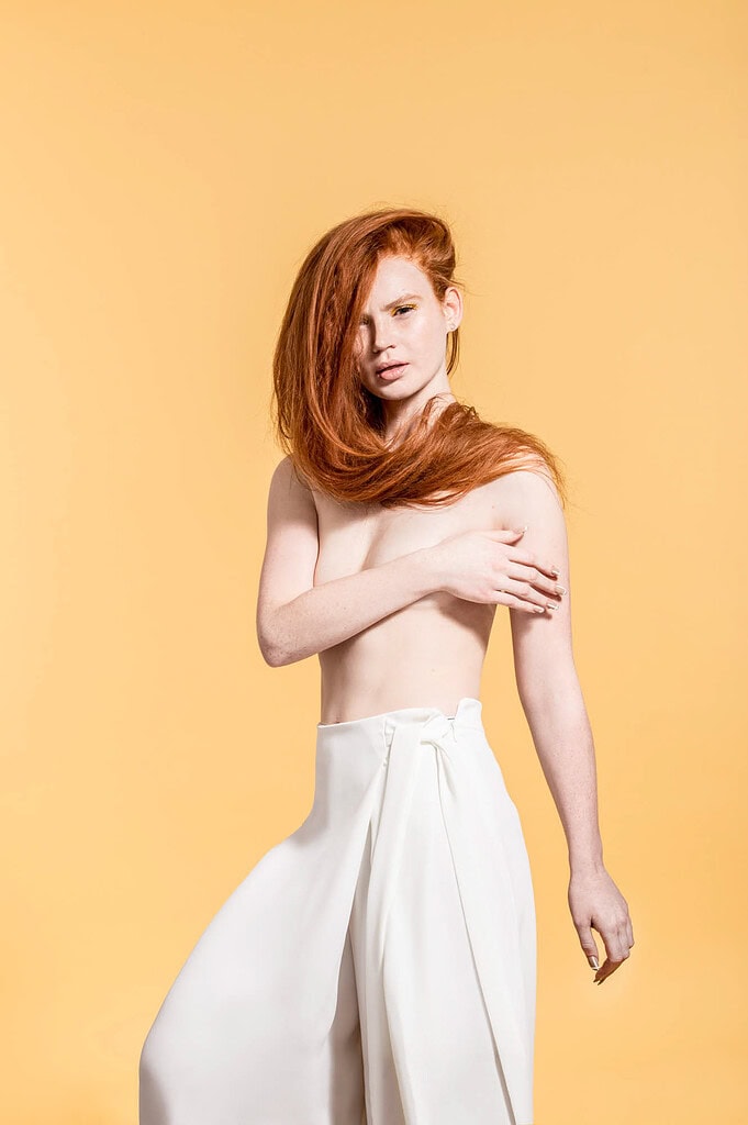 Red-haired model posing in white wide-leg trousers against a warm yellow studio background.