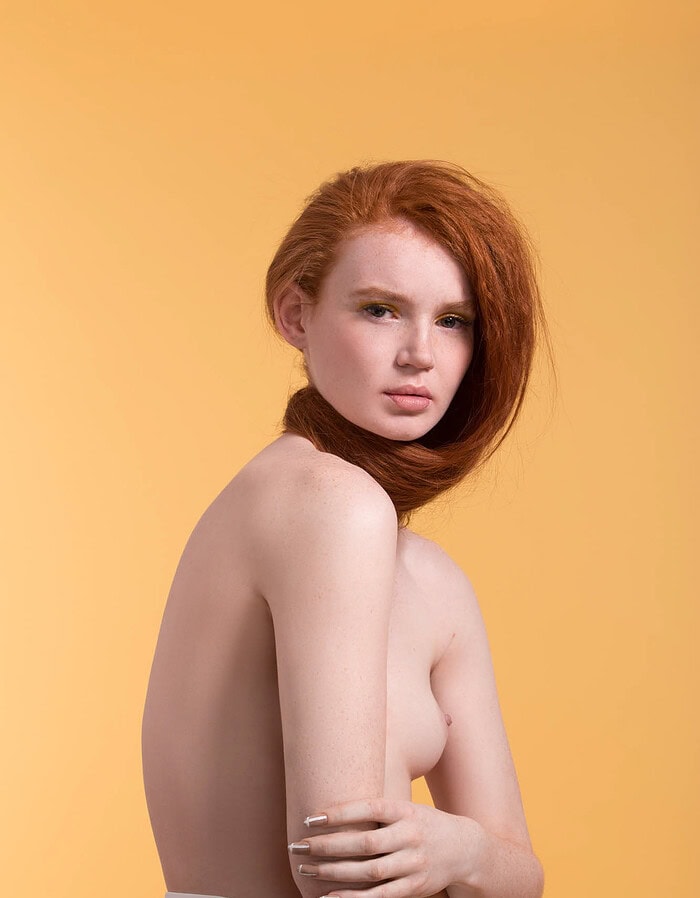Red-haired fashion model with her hair wrapped around her neck against a warm yellow studio background.