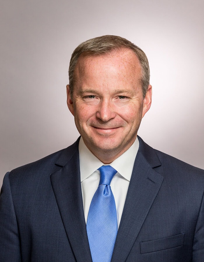 Male corporate professional wearing a dark suit and blue tie against a neutral studio background.