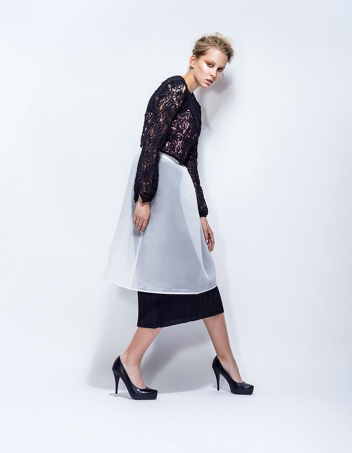 Model wearing a black lace top, layered black skirt and translucent white overskirt against a pale studio background.