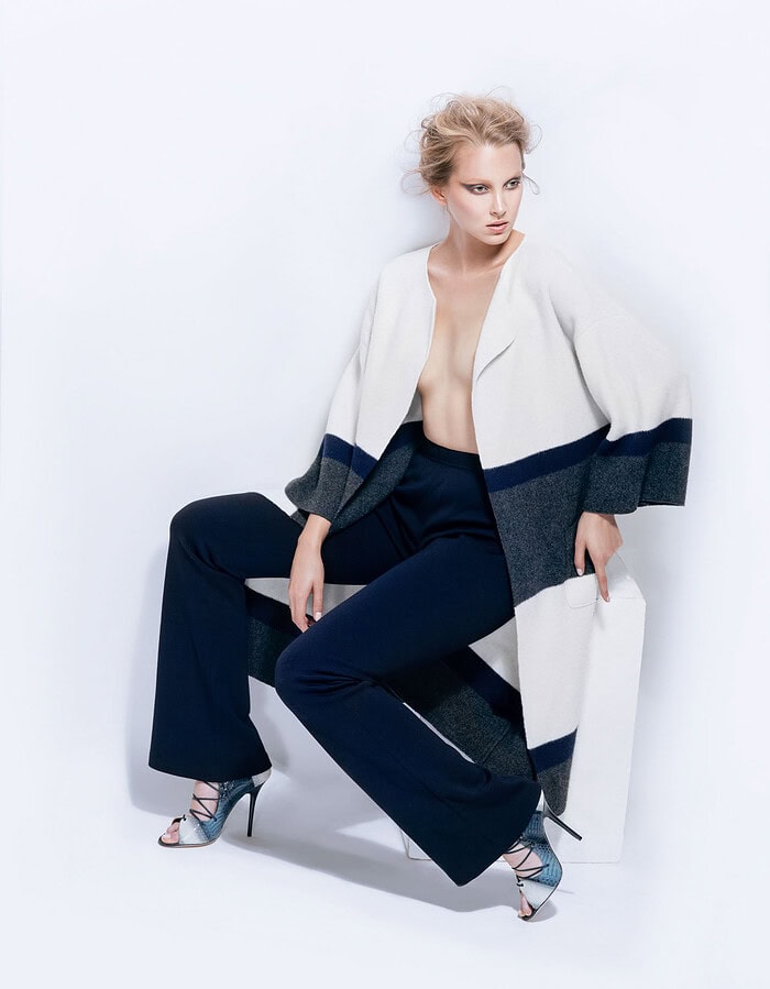 Model seated in navy flared trousers and a white, gray and blue oversized coat with metallic heels.