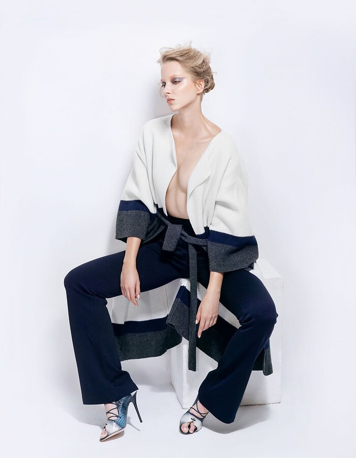 Model seated on a white block wearing a belted white, navy and gray coat with navy trousers.