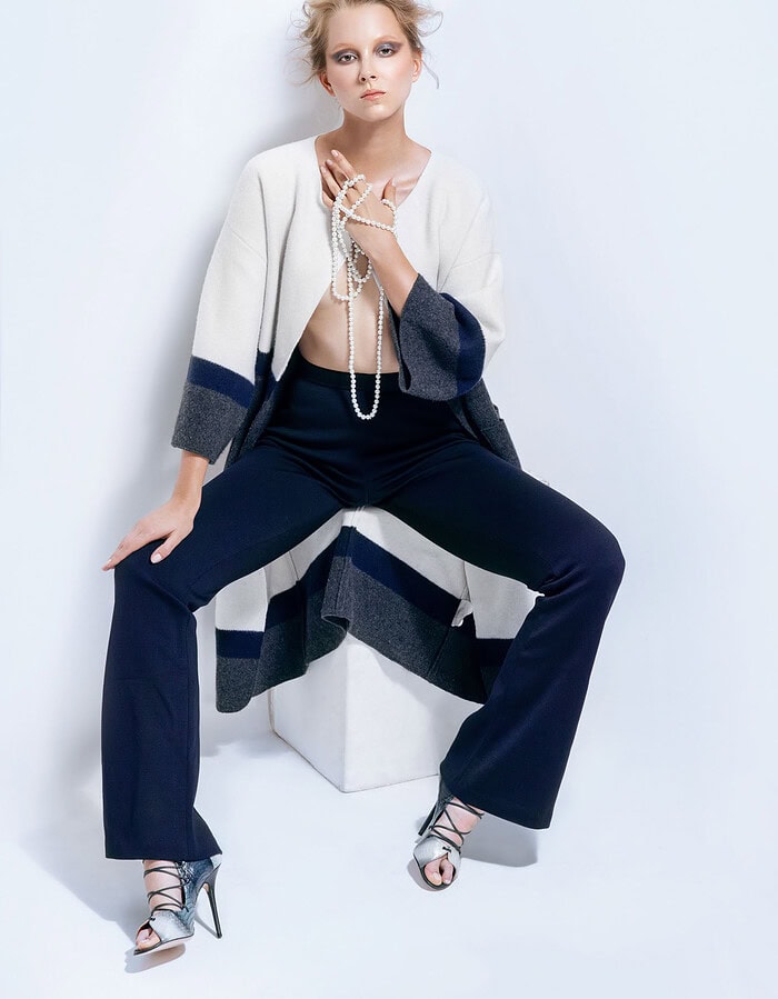 Model seated in navy trousers and a white color-blocked coat while holding long strands of pearls.