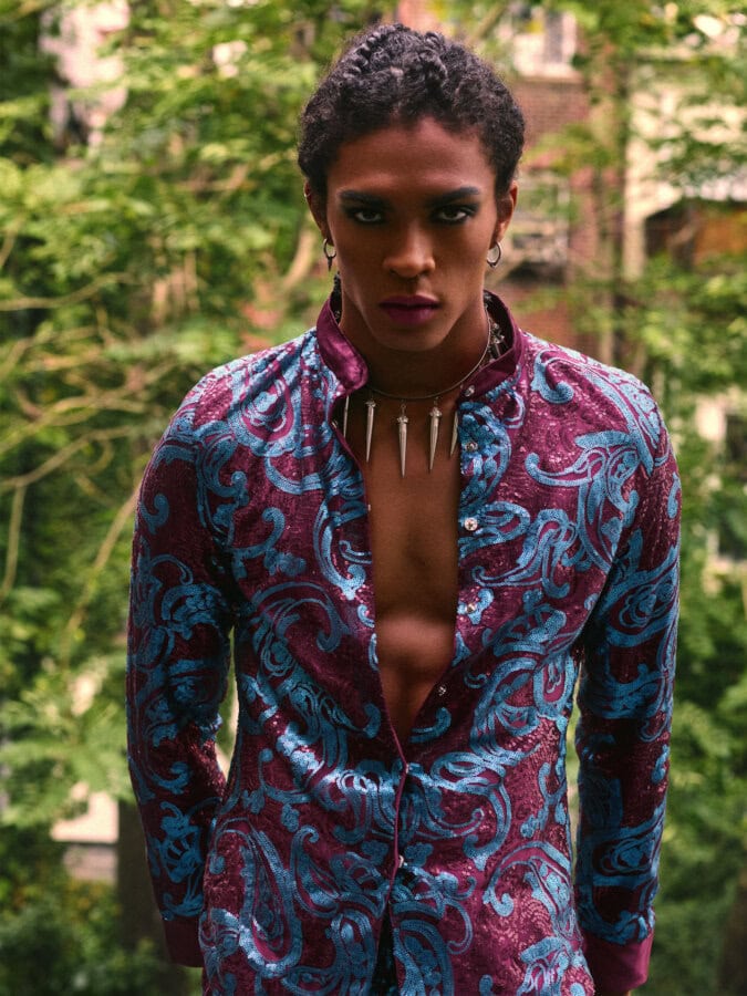 Menswear model wearing a burgundy and blue sequined jacket and pointed necklace in a green East Village garden.