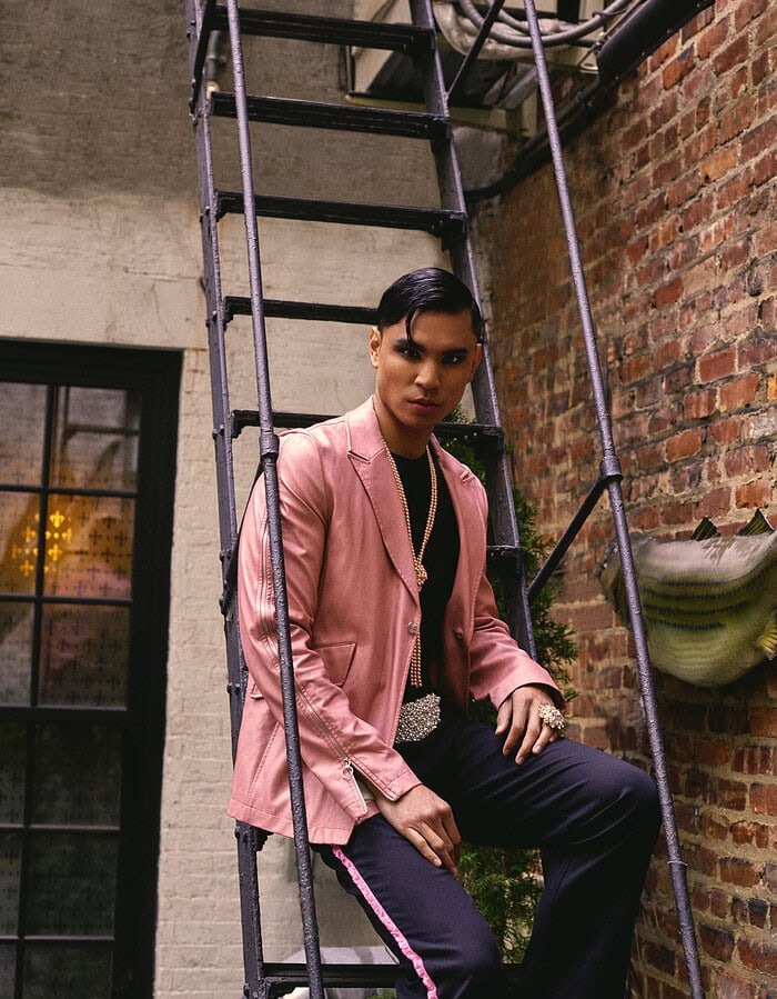 Model seated on an iron fire escape wearing a pink jacket, dark trousers and pearl jewelry.