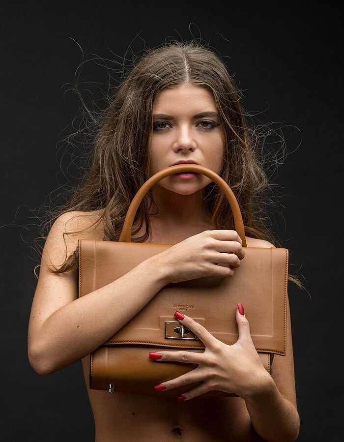 Beauty model holding a tan structured leather handbag against a black studio background.