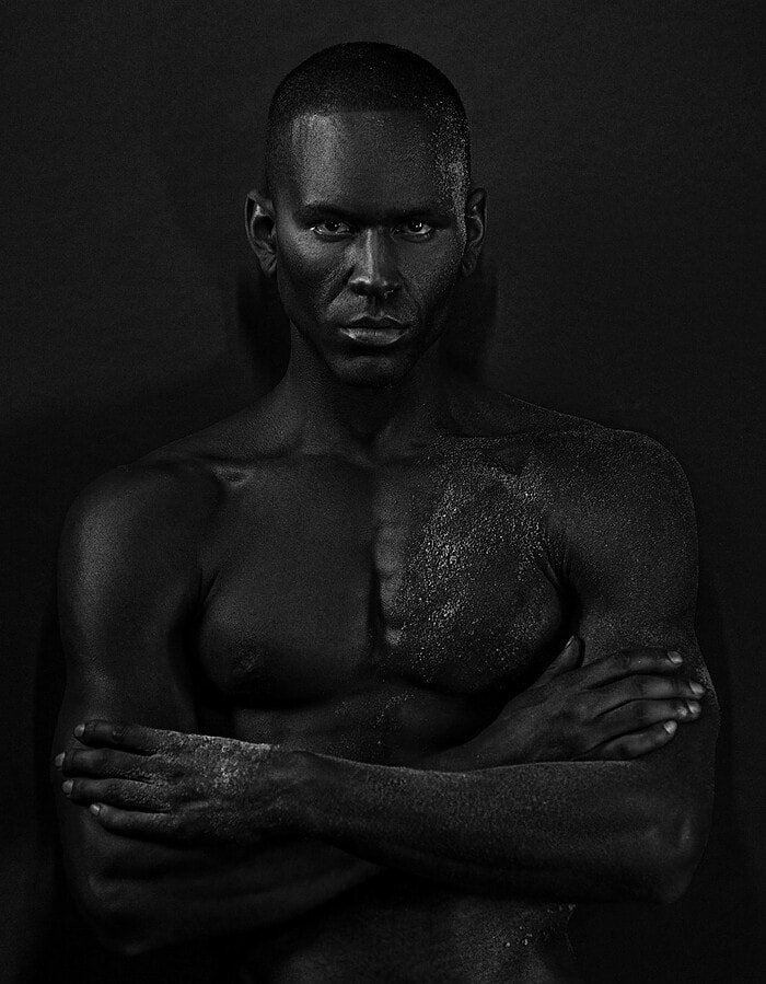 Male model standing with crossed arms in a dark black-and-white studio portrait