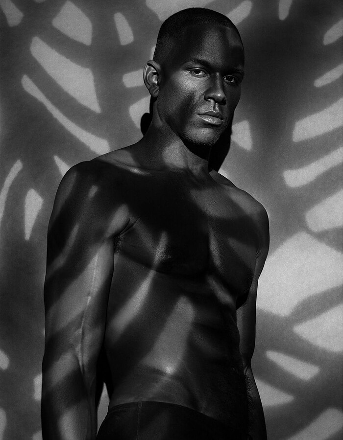 Black and white portrait of a muscular man with dramatic patterned shadows across his body and the background wall