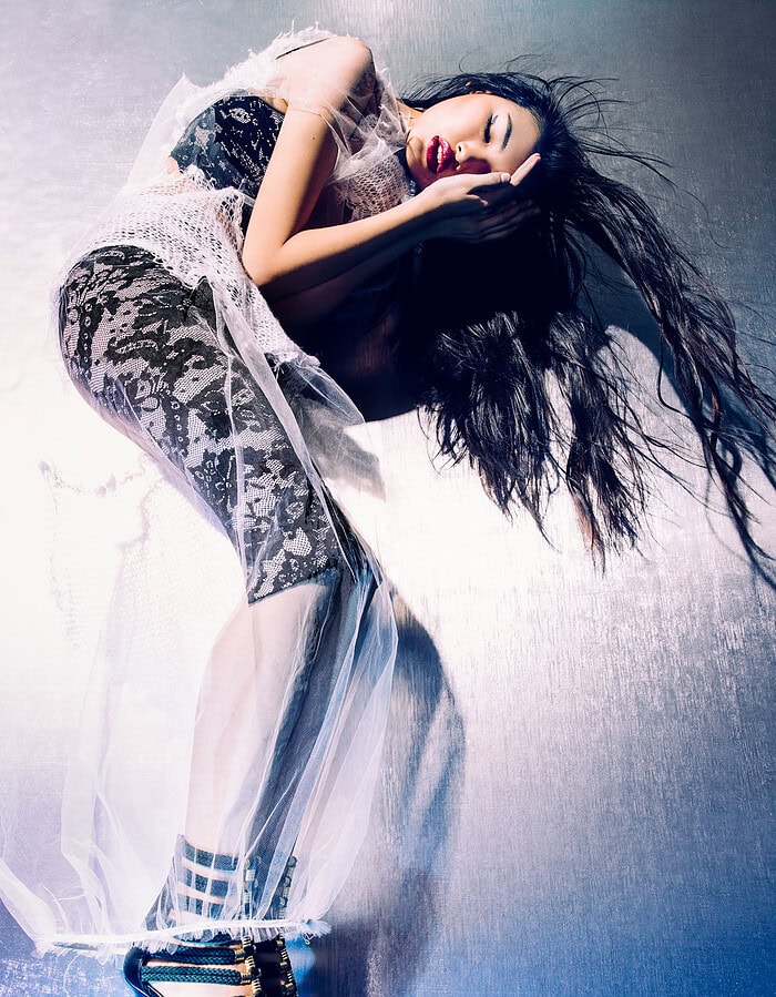 Fashion model bending sideways in black lace and sheer white tulle against a metallic blue wall.
