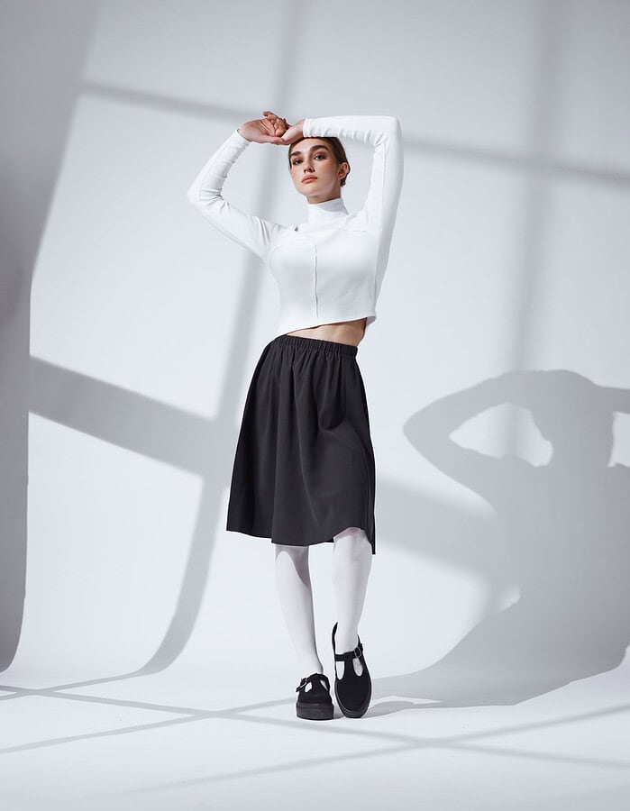 Fashion model in a fitted white crop top and black skirt posing with her arms raised beneath window shadows.