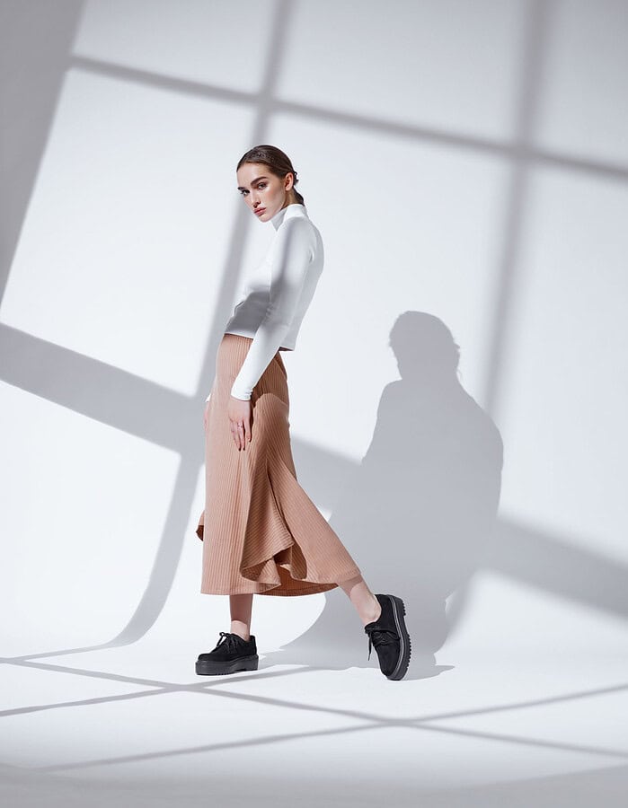 Fashion model walking in a white fitted top, tan pleated skirt, and black platform shoes.