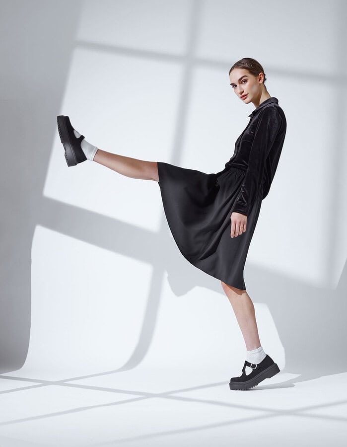 Fashion model in a black jacket and flowing black skirt posing with one leg extended.