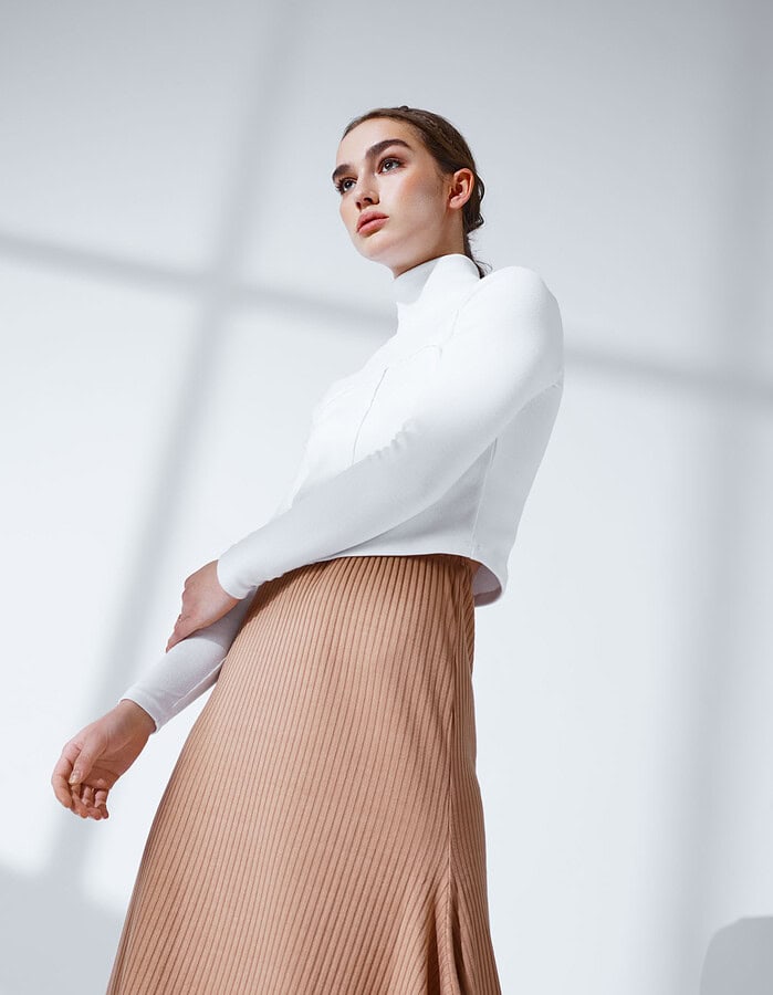 Low-angle portrait of a fashion model wearing a white turtleneck and tan pleated skirt.