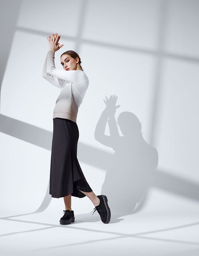 Fashion model in a light long-sleeved top and dark pleated skirt posing with raised hands.