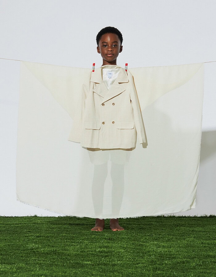 Child model standing behind translucent fabric with a cream blazer clipped across the front.