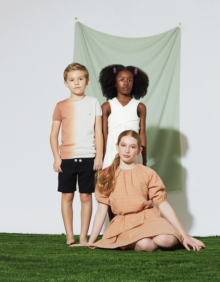Three child models pose in peach, white, and neutral outfits before a pale green fabric backdrop.