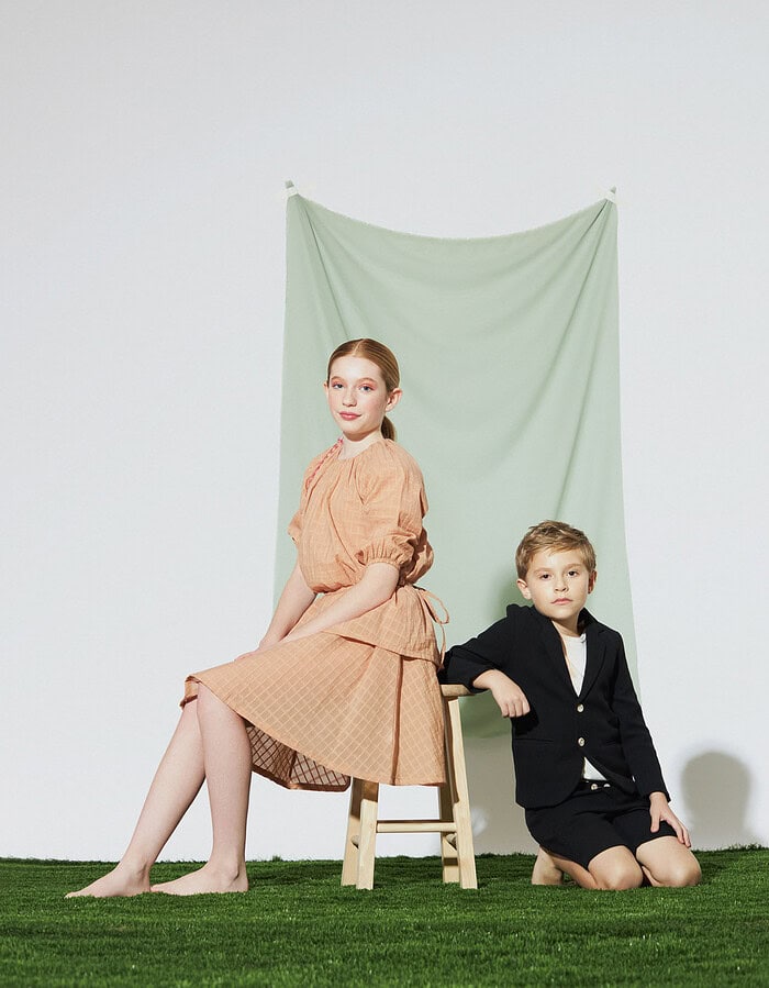 Two child models pose with a wooden stool in a peach dress and black tailored outfit.