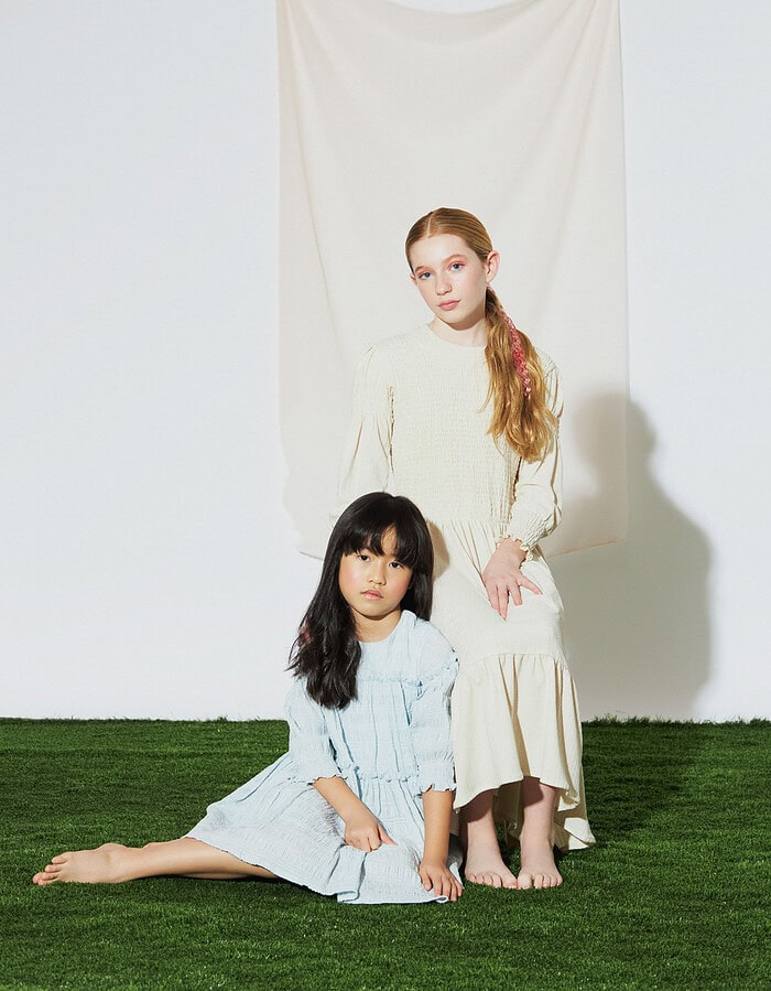 Two child models pose together in cream and pale blue dresses on artificial grass.