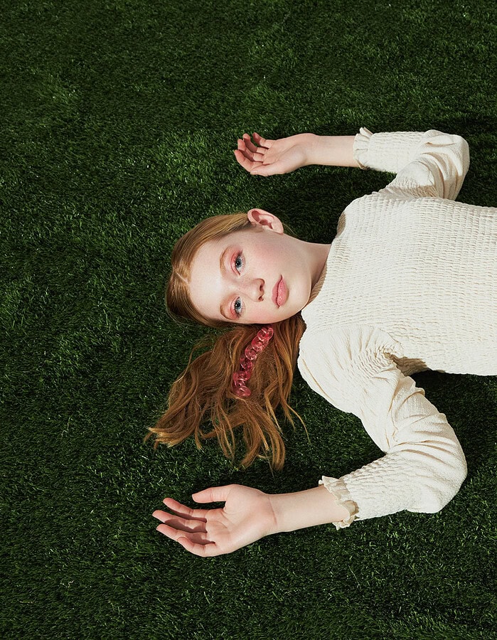 Child model in a long cream dress lies on artificial grass with her arms extended.