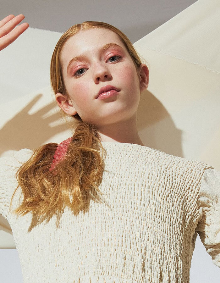 Close portrait of a child model in a textured cream dress with hands raised beside her face.