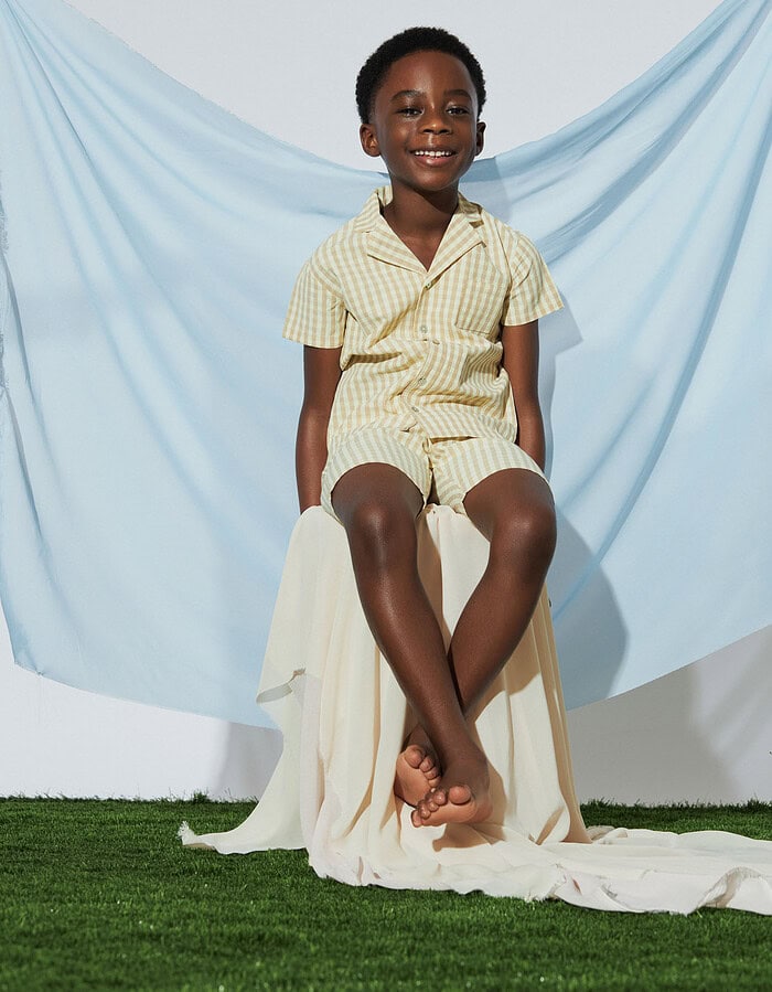 Smiling child model in a cream gingham shirt and shorts seated before pale blue fabric.