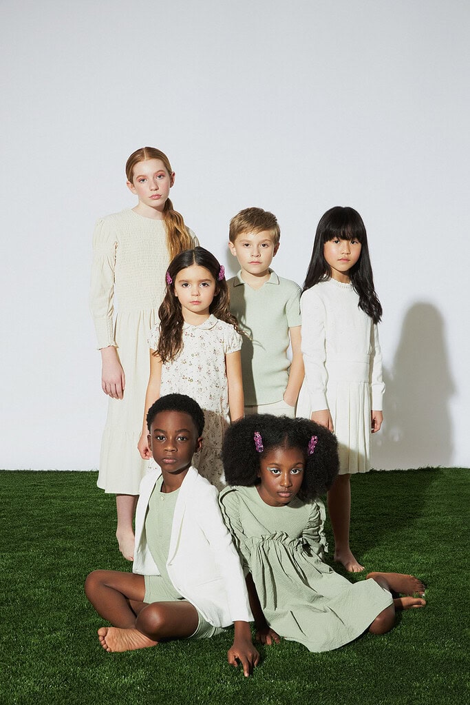 Six child models wearing cream and pale green outfits pose together on artificial grass.