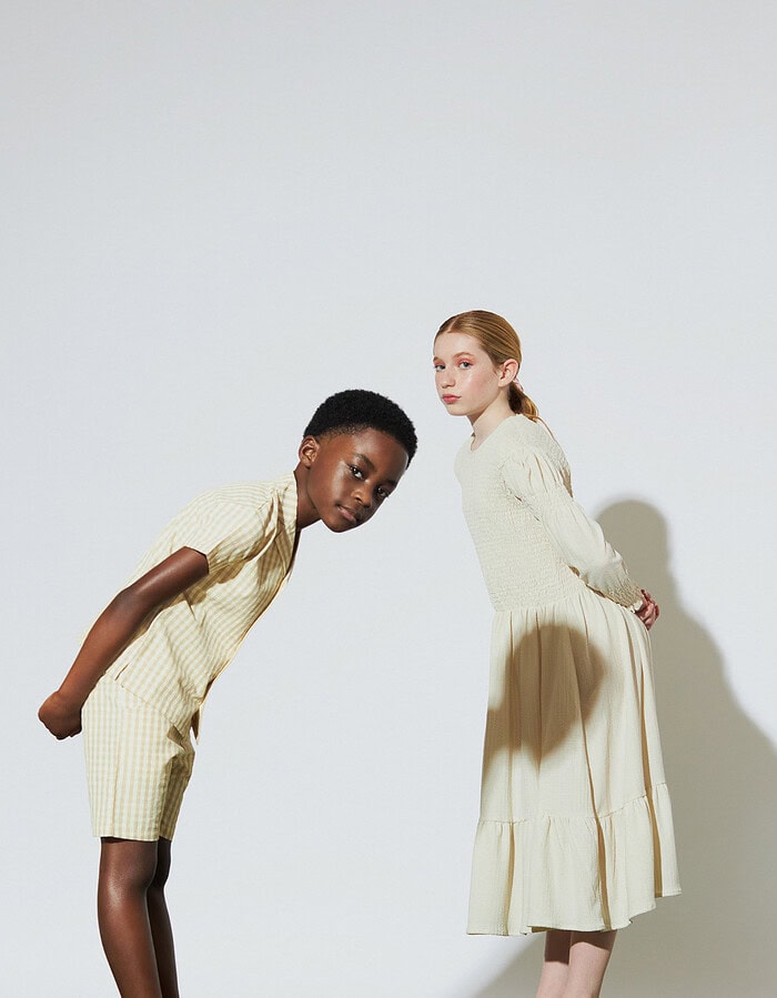 Two child models lean toward the camera in a gingham set and long cream dress.