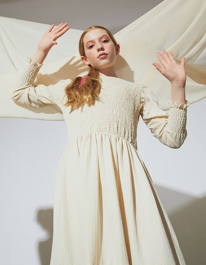 Low-angle portrait of a child model in a long cream dress with both hands raised.