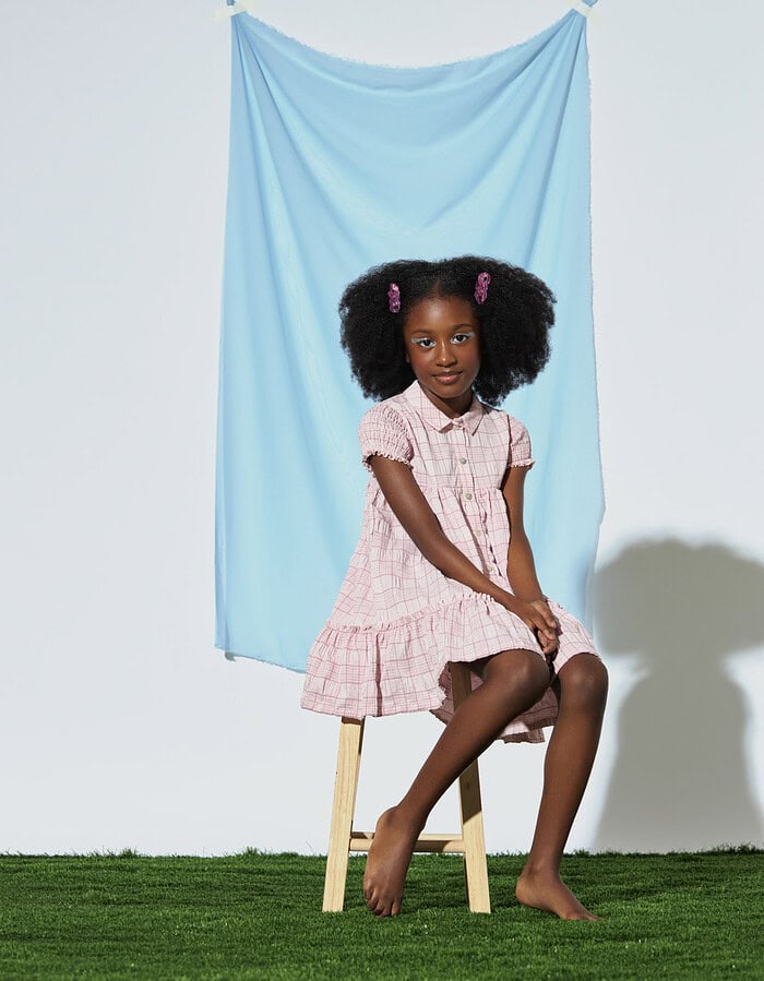 Child model in a pink checked dress seated on a wooden stool before blue fabric.