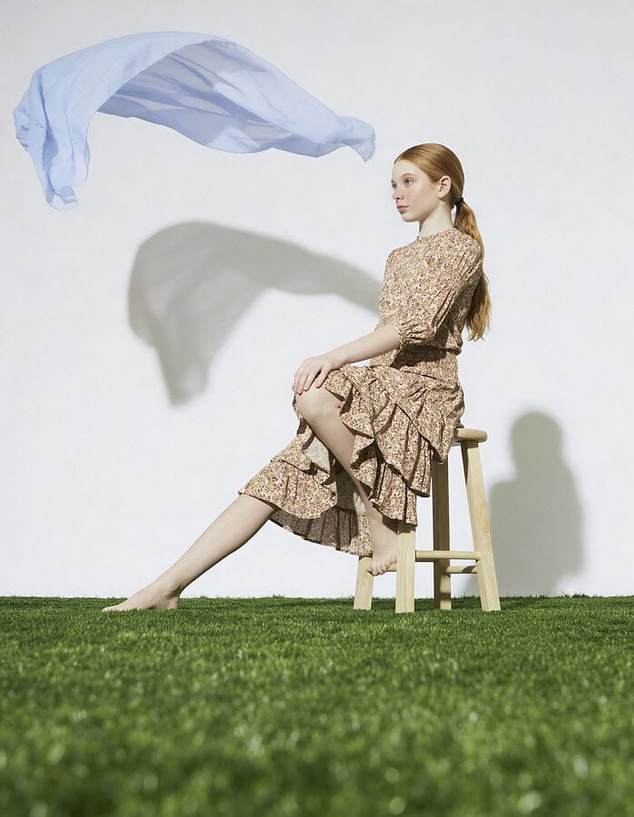 Child model in a floral dress sits in profile beneath floating pale blue fabric.