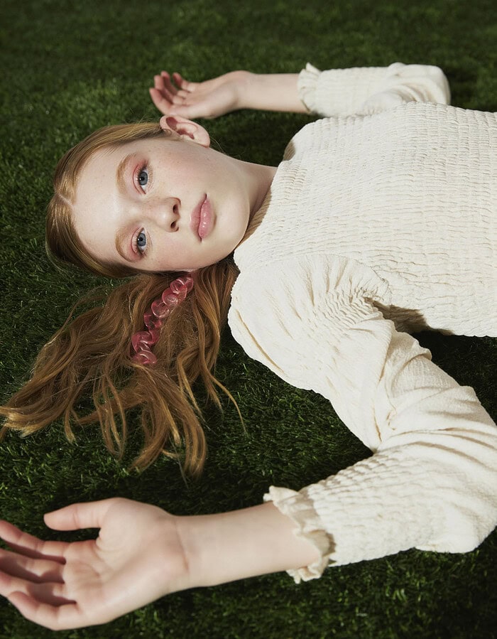 Close overhead portrait of a child model lying on grass in a textured cream dress.