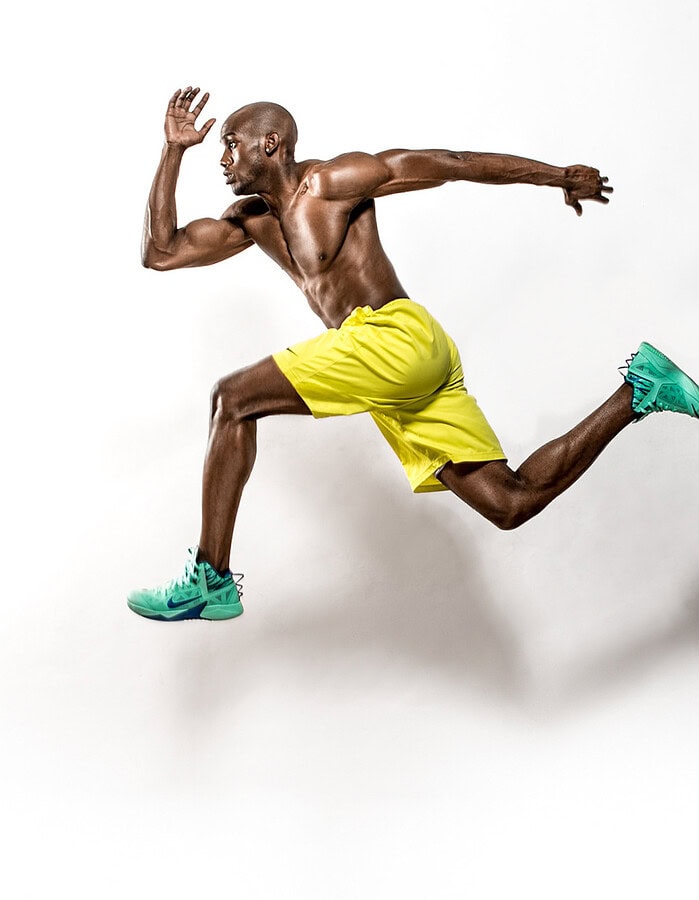Shirtless athletic model captured midair in yellow shorts and mint-green running shoes.