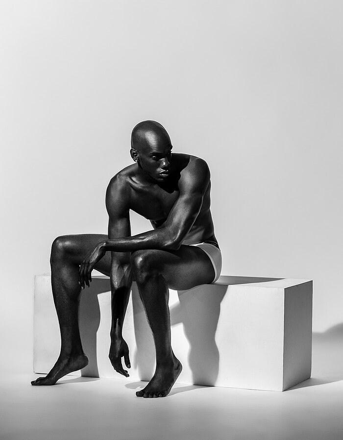 Black-and-white portrait of an athletic male model leaning forward while seated on a white block.