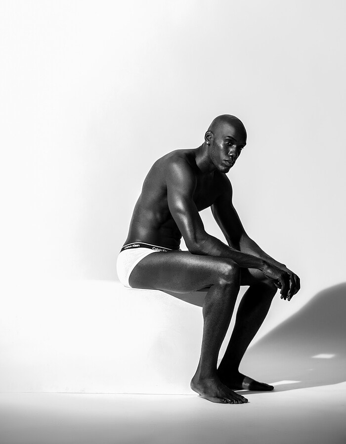 Athletic male model seated on a white block in a black-and-white studio portrait.
