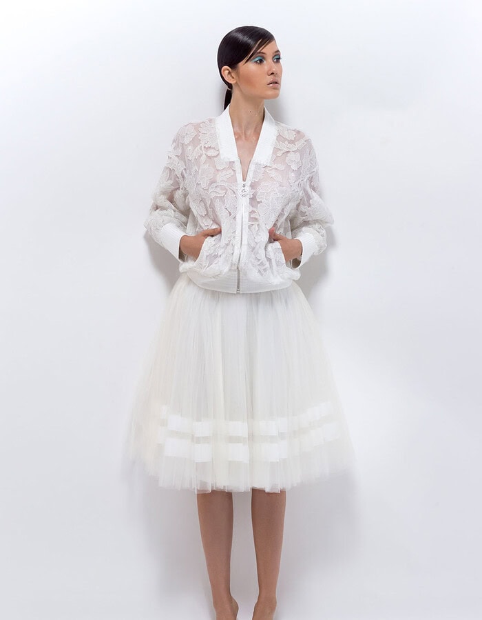 Full-length model in a white lace bomber jacket and knee-length tulle skirt against a white wall.