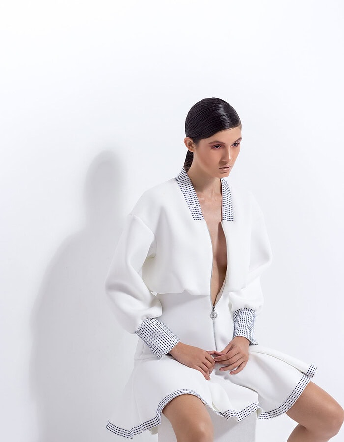 Model seated in a structured white dress beneath a large area of negative space in a white studio.