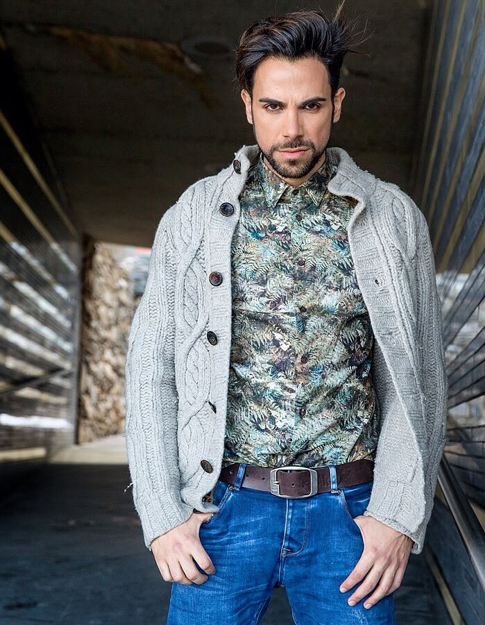 Portrait of a man in a patterned shirt and knit cardigan standing in a modern architectural corridor.