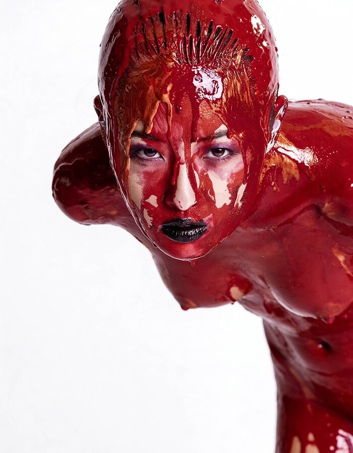 Person covered head to body in glossy red paint with black lips, leaning forward and staring intensely at the camera