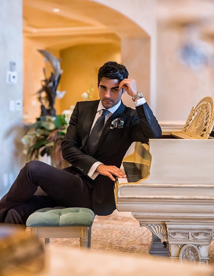 Formal portrait of a man in a dark suit seated at a white piano in an elegant interior.