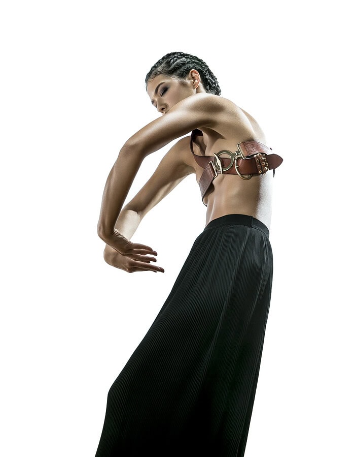 Fashion model bending her arms into a curved dance pose while wearing a leather belt top and black pleated trousers.