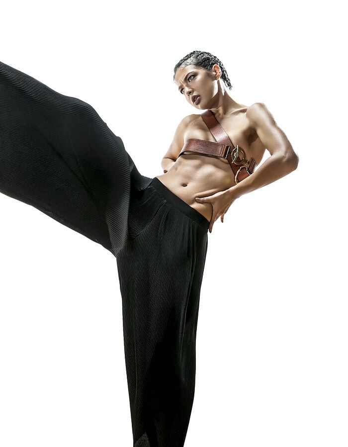 Fashion model in flowing black pleated trousers and a brown leather belt top performing an extended dance kick.