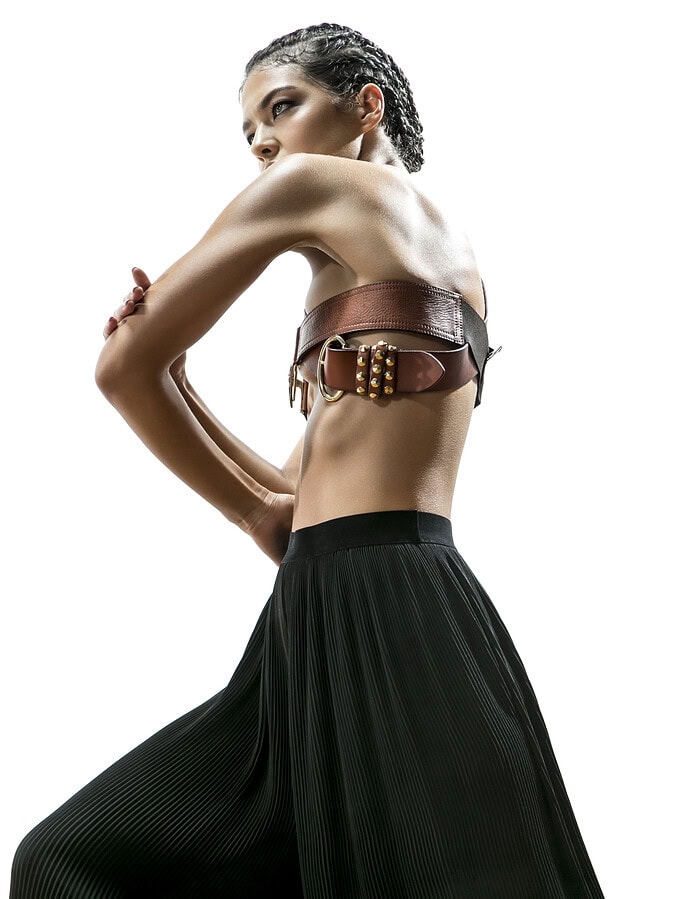 Fashion model viewed from behind wearing a brown leather belt top and flowing black pleated trousers.