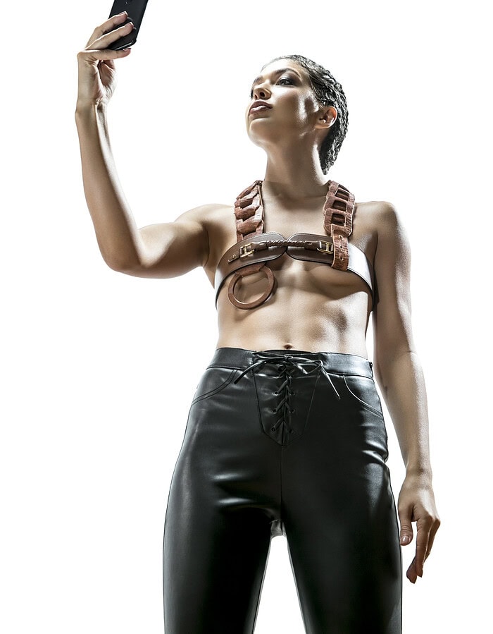 Fashion model holding a smartphone while wearing a leather belt top and high-waisted black trousers.