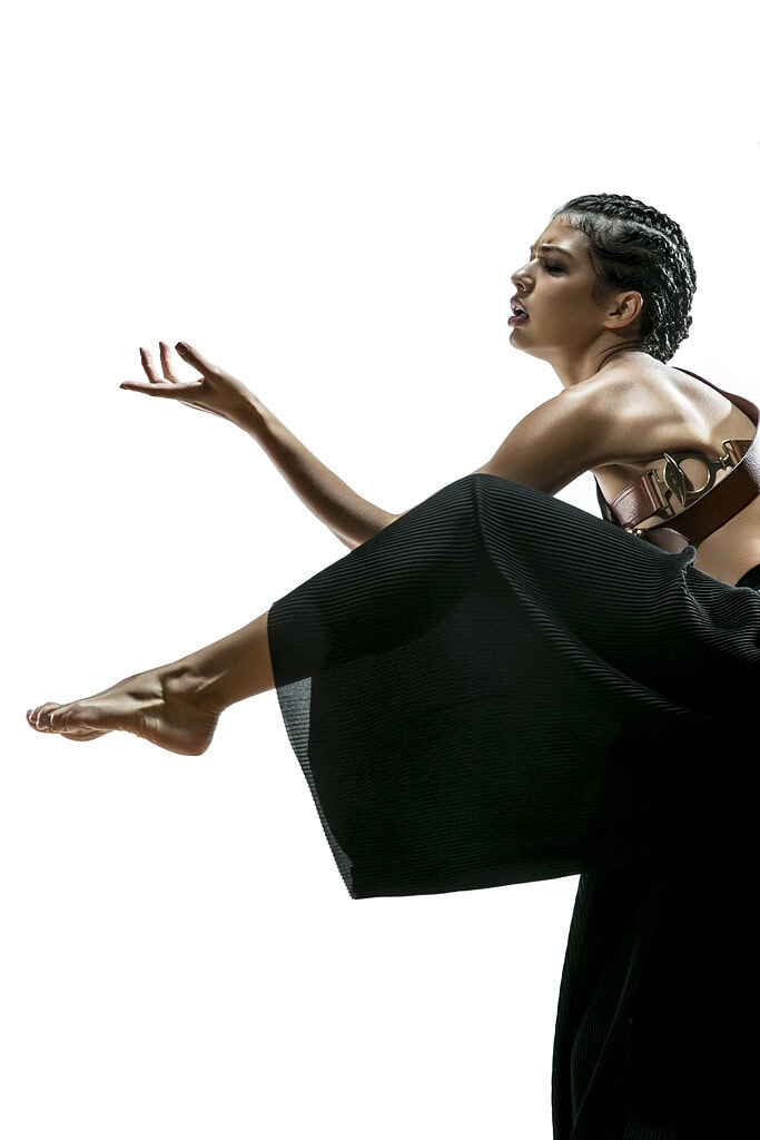 Fashion model in black pleated trousers posing with one leg extended and one hand raised.