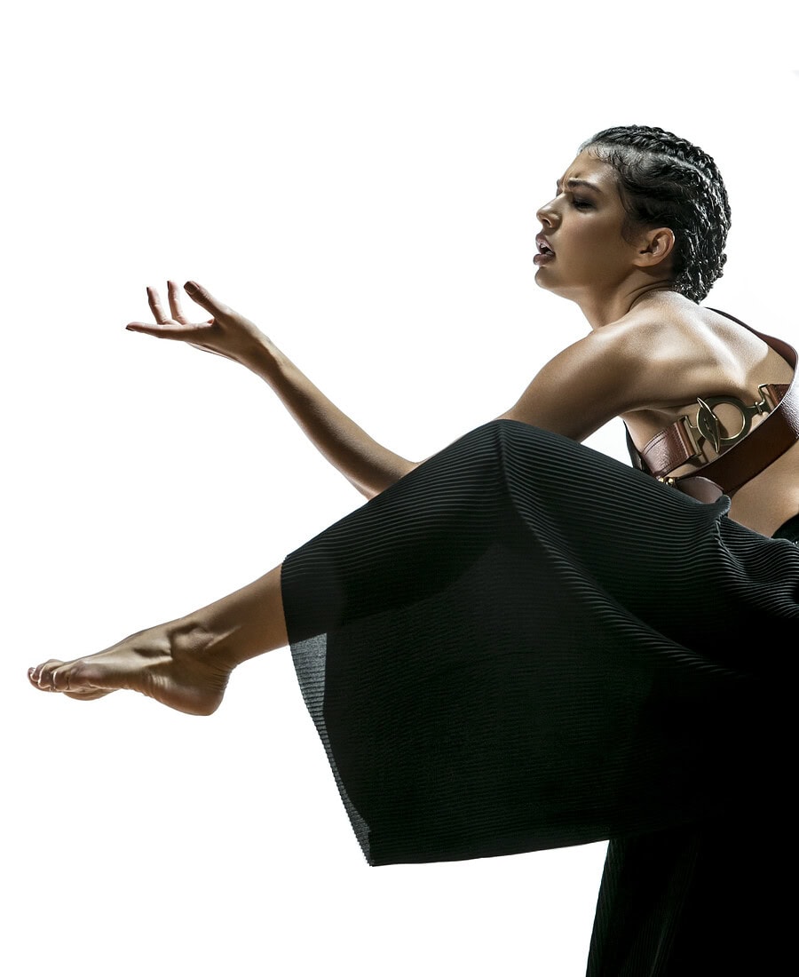 Fashion model in black pleated trousers posing with one leg extended and one hand raised.