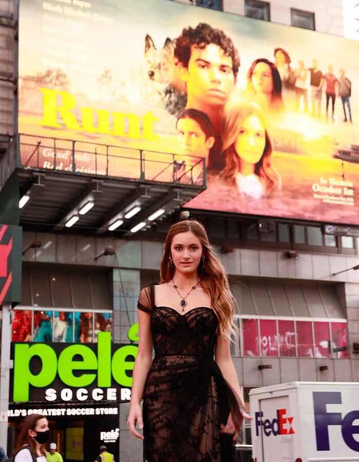 Music artist in a black dress photographed in Times Square beneath a large promotional billboard.
