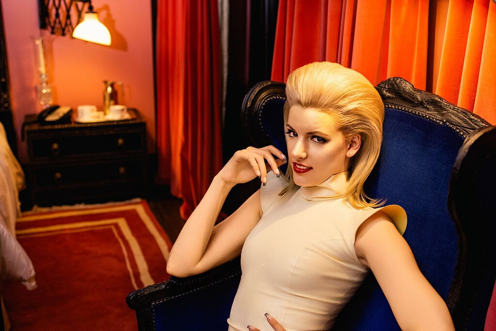 Music publicity portrait of a blonde woman reclining in a blue chair inside a richly colored room.
